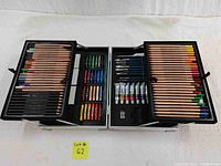Top-down view of open black carrying case filled with colored pencils, paintbrushes, oil pastels, acrylic paint tubes, and other drawing tools.