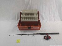 Fenwick Woodstream tackle box open showing three-tiered translucent trays, matched with Zebco retractable fishing pole laid beneath it on a white cloth background.