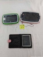 Photo showing three devices: Leapfrog LeapPad 3 (green and white), LeapPad Platinum e-reader (white and gray), and black touchscreen e-reader in pink case.