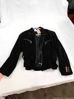 Black leather jacket laid flat on white surface, showing front with gold zippers and sleeve logo.