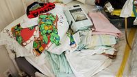 Pile of bed linens including sheets in pastel colors, full white/pastel bed skirt, Teenage Mutant Ninja Turtles themed sheets, and a Teenage Mutant Ninja Turtles knitted beanie on top.