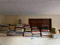 Wide view of multiple stacks of books placed on a high shelf, showing books of various sizes and colors including some hardcover and paperback items.