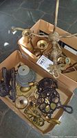 Two cardboard boxes filled with assorted brass and cast iron items including trivets, a bell, a lamp base, and candle holders.