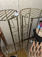 Two tall, cylindrical brass plant stands with round open rod tops and decorative swirl pattern legs standing on floor.