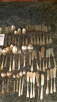 Top view photo showing rows of assorted silverplated forks, spoons, and butter knives laid out on a dark textured surface, highlighting the quantity and variety.