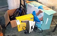 Photo showing boxed Homedics foot spa, Shark cordless vacuum inside a box, Black & Decker steam iron on the floor, and yellow Conair Pro blow dryer box underneath the iron.