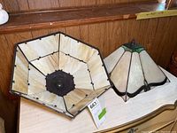View of two lampshades, one large hexagonal cream-colored slag glass and a smaller Tiffany style stained glass shade with cream and green glass panels.
