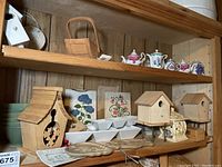Wide view of wooden shelves holding small ceramic miniature tea pots, wooden birdhouses, ceramic plaques with floral designs, small bowls, and decorative items.