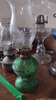 Four oil hurricane lamps, front left green pressed glass lamp with ornate details, metal burner and tall chimney, surrounded by three other lamps with varied bases and clear chimneys.