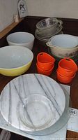 Photo showing Pyrex Homestead pattern casserole dish with lid, yellow and white milk glass mixing bowls, clear glass mixing bowls stacked, orange Rachael Ray single-serve bowls, marble lazy Susan on the wooden surface.