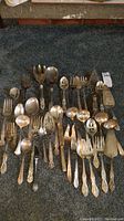 Top-down view of assorted silver plated and stainless steel cutlery and serving utensils laid on a dark carpet, showing a variety of forks, spoons, and pie servers with ornate handles and some tarnish.