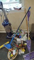 Shark upright vacuum cleaner on left with clear canister, surrounded by assorted cleaning supplies and tools on floor near wall