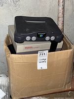 Photo showing a vintage Nintendo 64 console sitting on top of a Nintendo Entertainment System console, both inside a cardboard box in a basement with rough wall and pipe in background.