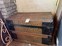 Closed vintage trunk with patterned exterior, wooden slats, black metal edges and corners, and visible wear.
