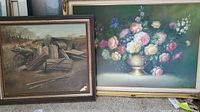 Both framed paintings leaning against wall: floral still life in gold-tone frame, landscape in brown frame.
