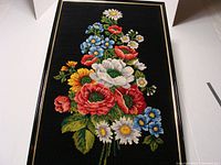 Full view of the framed floral petit point needlepoint artwork with a black background and a bouquet of various flowers including red poppies, white daisies, and blue forget-me-nots.