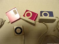 Four Apple iPod Mini devices in different colors with three white charging docks connected to USB cables, some devices positioned in docks, one powered on.
