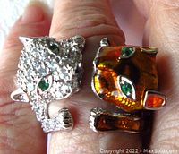 Close-up of ring showing two feline heads with green eyes, clear stones on one head and orange enamel on the other.