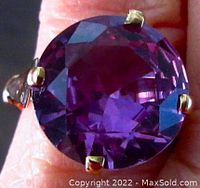 Close-up front view of faceted round purple stone set in yellow gold ring on finger, shows color and prong setting detail.