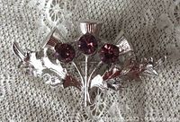 Front view of the silver-tone brooch showing three purple glass stones and two leaves, laying on lace fabric.