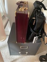 Side view of briefcases and filing cabinet stacked, showing burgundy Franzen briefcase and black Billblass attaché with metal cabinet below
