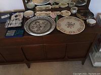 Overview of lot on wooden sideboard showing large platters, figurines, plates, and coasters.