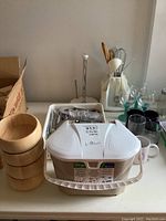 Photo showing stacked wooden salad bowls, white compost bin, packaged cutlery set, utensil caddy, paper towel holder, assorted glassware and mugs on a countertop.