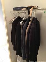 Several men's jackets hanging on a closet rod, dark colors including black and navy, with a belt on top shelf and a Tommy Hilfiger box visible.