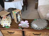 Photo of four items on a wooden dresser: vintage lidded Italian candy bowl, boxed Asian vase, pink petal-shaped bowl, and ornate mirrored tray.