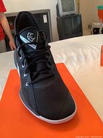 Front view of black Nike athletic shoe on an orange box showing mesh upper, laces, and tongue logo.