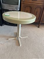 Round metal outdoor table with light green tripod base on carpeted floor, showing side view.