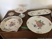 Five porcelain serving pieces on brown surface including large pierced plate, pedestal dessert stand, small round dish, elongated serving platter, and large flat plate all decorated with floral motifs and gold trim