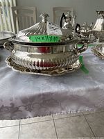 Silver plated tureen with lid and tray on a table with a white patterned tablecloth, note of dimensions on green tape visible.
