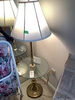 Table lamp with metal base and clear glass section, topped with white lampshade, shown on a side table near a floral chair and a wall outlet.