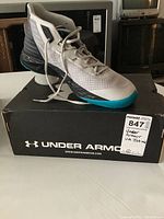 Side view of left shoe placed on top of original Under Armour shoe box, showing mesh upper, lacing, and toe area with turquoise outsole edges.