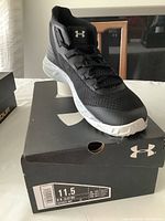 Angled front view of one black Under Armour UA Jet Mid athletic shoe sitting on the original shoe box with size label 11.5 visible.