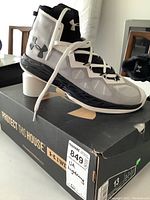 Side view of white and black Under Armour UA Lightning 4 shoe on top of its box showing branding and size details.