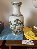 Full front view showing full form and decoration of the white porcelain vase with hand painted Chinese landscape and trees, including decorative black borders near rim and base. The vase is placed on a blue cloth with part of a yellow cloth and a lamp nearby.