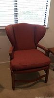 Front view of upholstered reddish-brown wingback armchair with shaped wooden legs and armrests under window blinds.