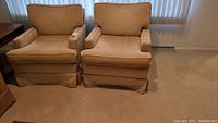 Two tan fabric arm chairs side by side on carpeted floor near window with vertical blinds. Both chairs have skirted bottom and appear slightly worn requiring cleaning.