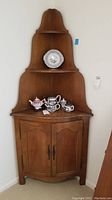 Front view of wooden corner hutch showing three shelves with decorative items and curved double doors below.