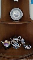 First image showing overall display of four teapots and one decorative plate on wooden shelf with the plate on upper shelf on stand and teapots on lower shelf.