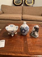 Photo showing the three items on a wooden table in front of a beige couch: a floral bowl on feet, a blue and white vase, and a figurine of a woman with a cow.