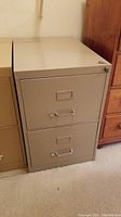 Beige metal filing cabinet with two drawers, handles, and lock on the top drawer. Shows surface chipping and wear.