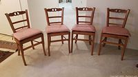 Four wooden dining chairs with ladderback design and pink upholstered cushions, arranged side by side on carpet.
