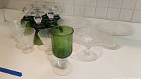 Photo of 8 green stained glass goblets with clear stems at front, 3 clear stemmed glasses, and 3 clear stemmed dessert dishes on kitchen counter.