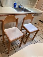 Two matching wooden kitchen stools with white leather-like padded seats placed in front of a kitchen counter and sink.