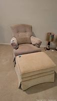 Beige fabric upholstered rocking chair with matching ottoman positioned in a room with a lamp and beige carpet. Chair has tufted back, padded arms, and lumbar pillow. Ottoman has a skirt and casters.