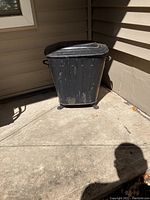 Photo of a black rectangular outdoor waste can with lid, showing surface wear and positioned against a beige wall on a concrete porch.