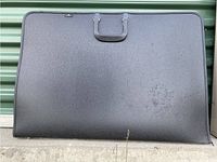 Closed view of the black textured artist's portfolio case resting against a green wall, shows size and handle.